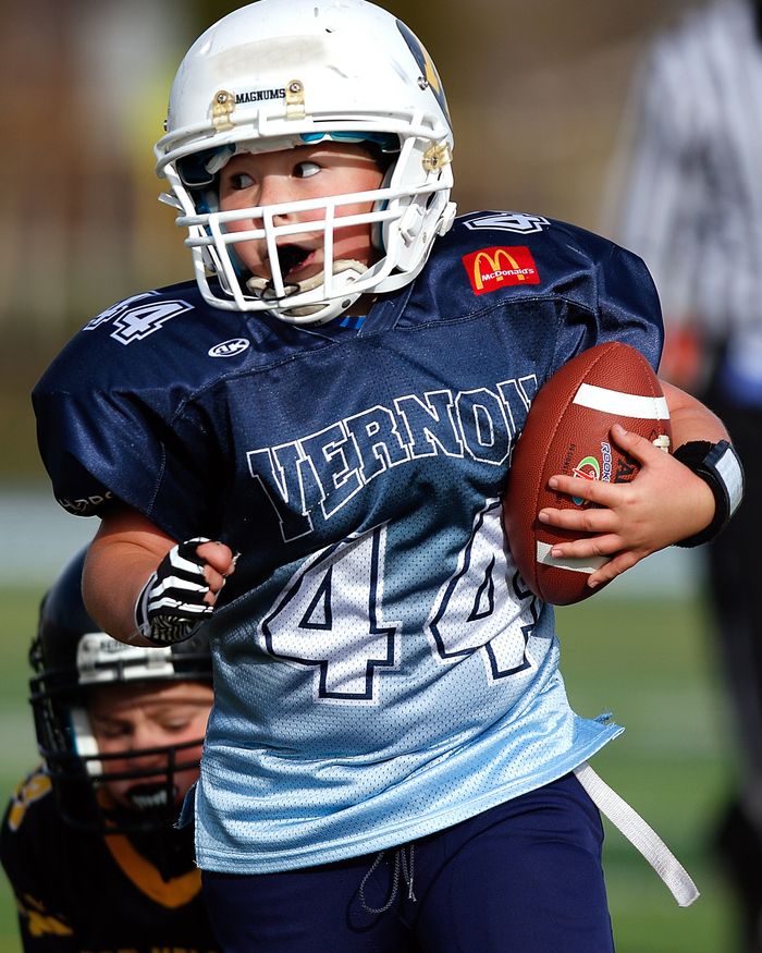A young child in football gear holding a ball during a game on the field.